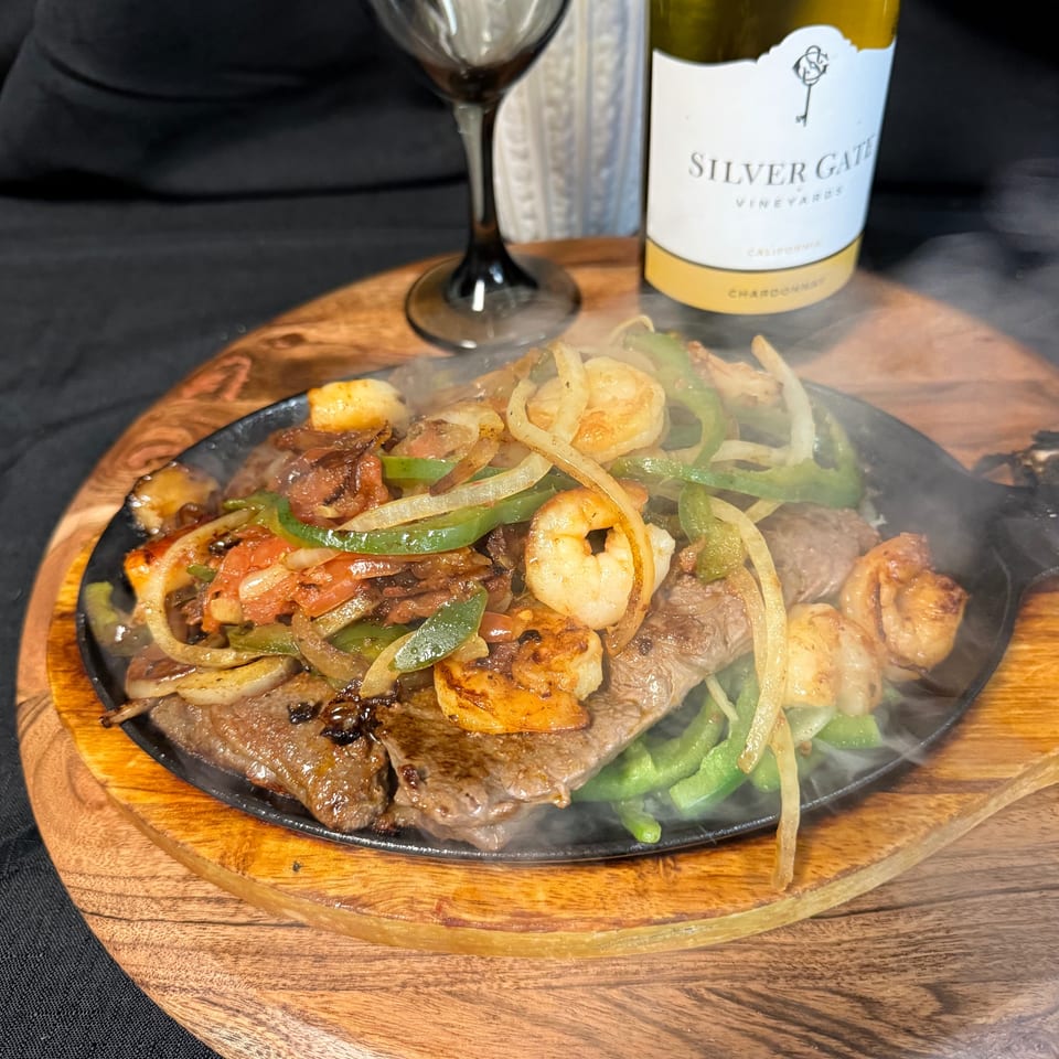 Sizzling steak and shrimp fajitas with bell peppers and onions on a wooden platter, with wine.