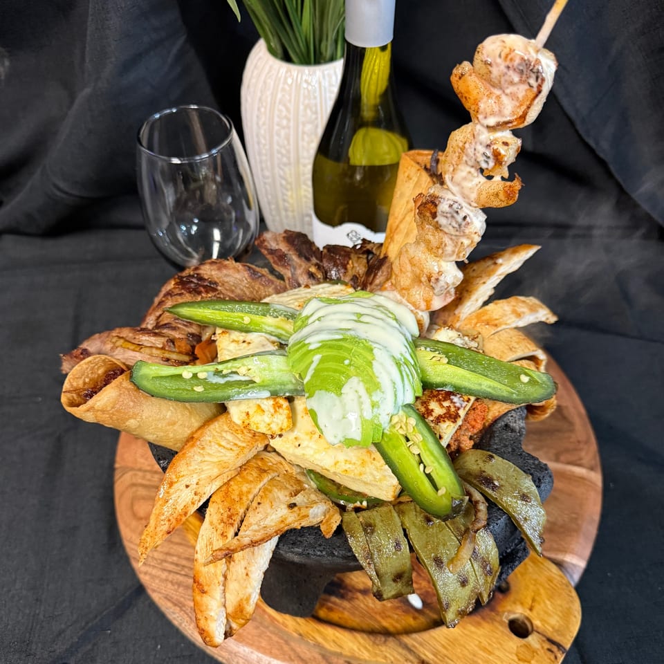 A large Mexican molcajete dish with grilled meats, cheese, avocado, jalapeños, and a shrimp skewer.